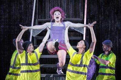 WNO Candide Soraya Mafi Cunégonde photo credit Craig Fuller 1790 Cunégonde sings out to the audience as she is lifted by two dancers dressed in hi-vis shirts and beanie hats, each with a cigarette in their mouths. She wears a corset top, denim shorts and a sparkly chain dress.