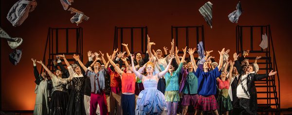 WNO Candide cast of Candide photo credit Craig Fuller 1159 The Old Woman stands centre stage, her arms in the air triumphantly. Behind her, the dancers and the WNO Chorus all do the same gesture, smiling gleefully. They have thrown multiple items of clothing into the air, which fly above the crowd.