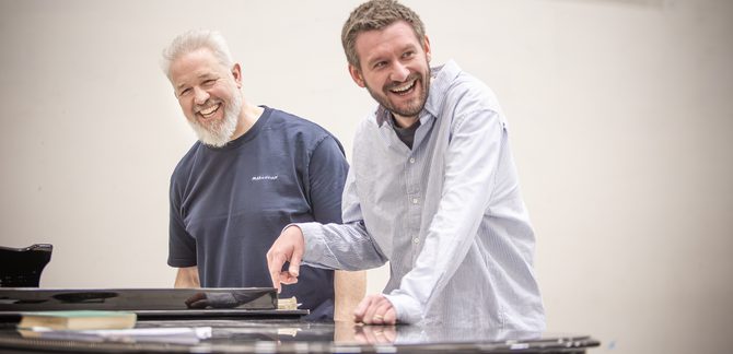 WNO The Flying Dutchman Simon Bailey The Dutchman Jack Furness Director photo credit Craig Fuller 4120 Simon Bailey and Director Jack Furness stand together behind a piano, smiling. Jack leans on the piano with one hand and points towards a book with the other.
