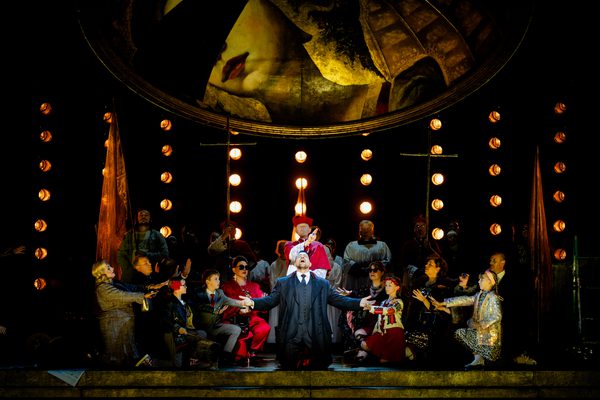 WNO Tosca Cast of Tosca Photo Credit Dafydd Owen 1004 Scarpia is on his knees at the centre of the stage. He wears a suit and formal jacket and raises his face and palms to the sky. Behind him is a priest, and a crowd of fashionable people and choral singers surround him, their arms reaching towards him. Many spotlights glow with light, and the golden dome hangs above.