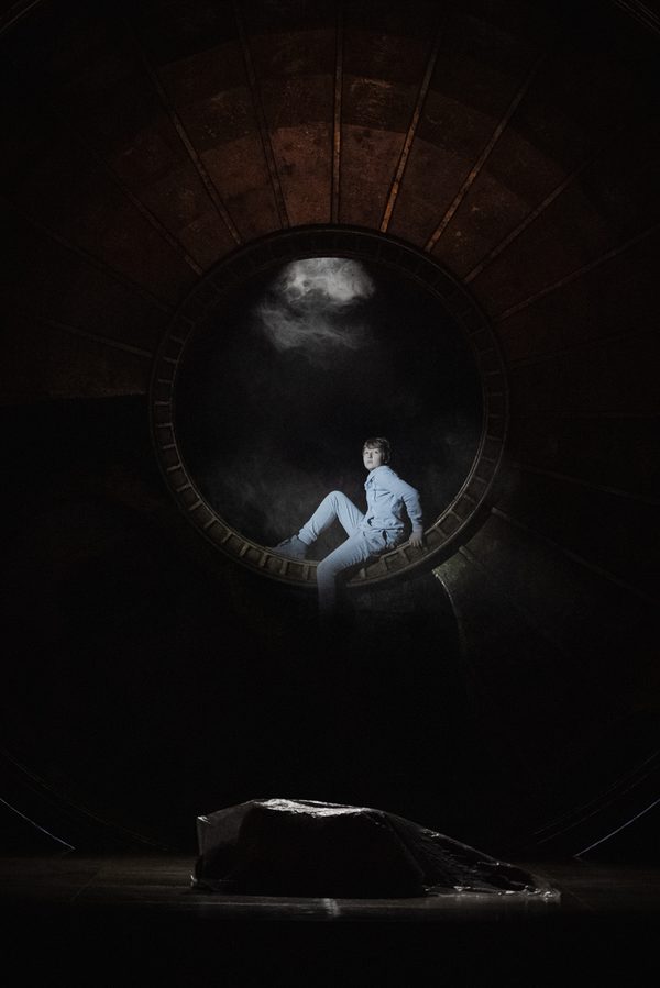 WNO Tosca Cast of Tosca Photo Credit Dafydd Owen 2063 A young boy in a hoodie and jogging bottoms sits at the centre of the golden dome in semi-darkness, a spotlight shining on him. On the ground is a body covered by a large plastic sheet.