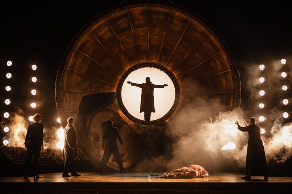 WNO Tosca Cast of Tosca Photo Credit Dafydd Owen 880 Tosca stands at the centre of the golden dome with her arms outstretched. Only her silhouette is visible against bright lighting. Below, Cavaradossi lies dead on the ground and various people look up at her.