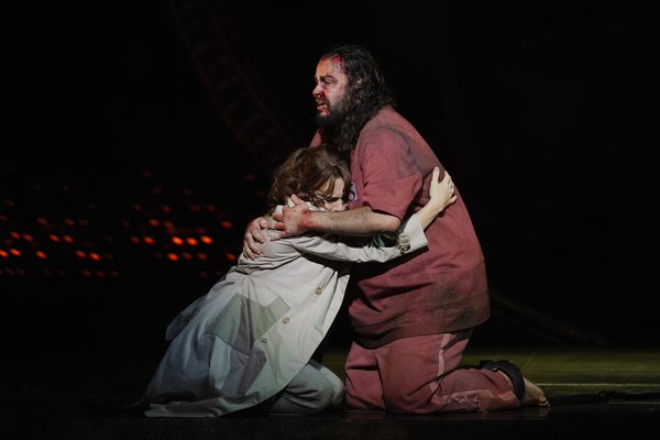 WNO Tosca Natalya Romaniw Tosca Andrés Presno Cavaradossi Photo Credit Dafydd Owen 2455 Tosca and Cavaradossi hold each other close. Cavaradossi wears a prisoner's outfit and is covered in blood and dirt. Tosca is on her knees and presses her head against Cavaradossi's chest.