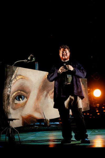 WNO Tosca Ross Fettes The Sacristan Photo Credit Dafydd Owen 589 The Sacristan stands facing the audience with a grin on his face, holding a vial of alcohol. Behind him is the fresco that Cavaradossi has been painting.