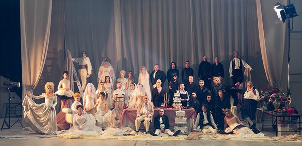 WNO 2627 Launch BCOR Cover Two groups of people - dressed in black and white are on a large stage. With a large velvet curtain behind them. One half is wearing wedding dresses - whilst the other is wearing sombre suits. In the middle, is a large table, with an array of wedding cakes and candles.