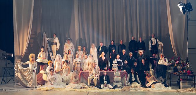 WNO 2627 Launch BCOR Cover Two groups of people - dressed in black and white are on a large stage. With a large velvet curtain behind them. One half is wearing wedding dresses - whilst the other is wearing sombre suits. In the middle, is a large table, with an array of wedding cakes and candles.