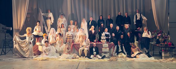 WNO 2627 Launch BCOR Header Two groups of people - dressed in black and white are on a large stage. With a large velvet curtain behind them. One half is wearing wedding dresses - whilst the other is wearing sombre suits. In the middle, is a large table, with an array of wedding cakes and candles.