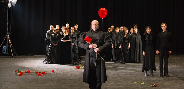 WNO 2627 Launch LB Cover A large group of people all dressed in black stand in the background. In the foreground, two lovers, and a man holding a red balloon and flowers
